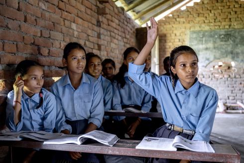 Elever iført blå skjorter sidder pænt på skolebænken og rækker hånden op