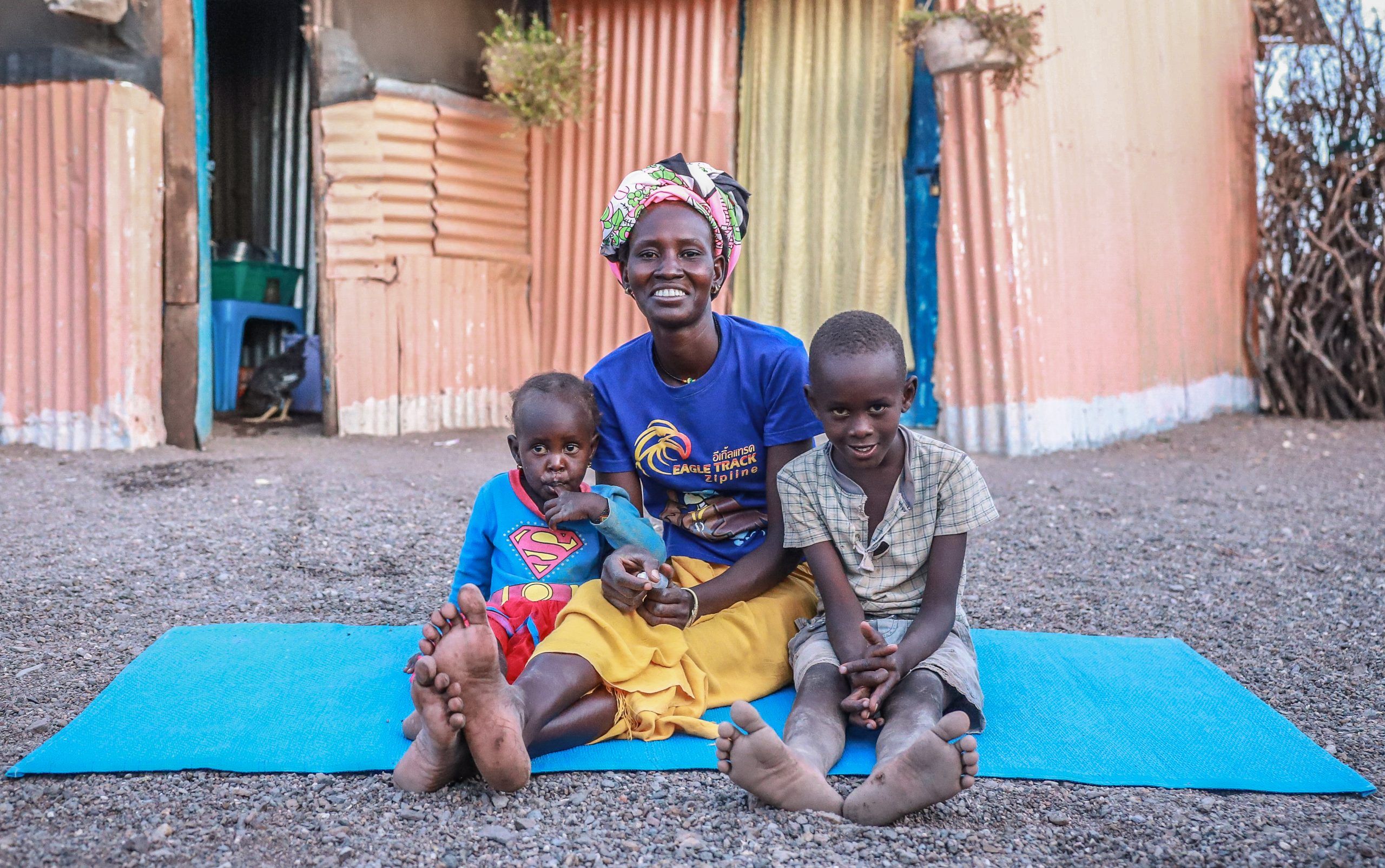 Emily sitting on a blue mat with her two children by her side smiling at the camera. Illustration: Danmarks Indsamling 2023.