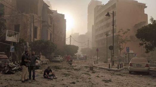 En såret mand sidder midt på gaden, mens to andre står og taler ved siden af. Skilte er ødelagt og grene og møbler ligger spredt ud over gader og fortove.