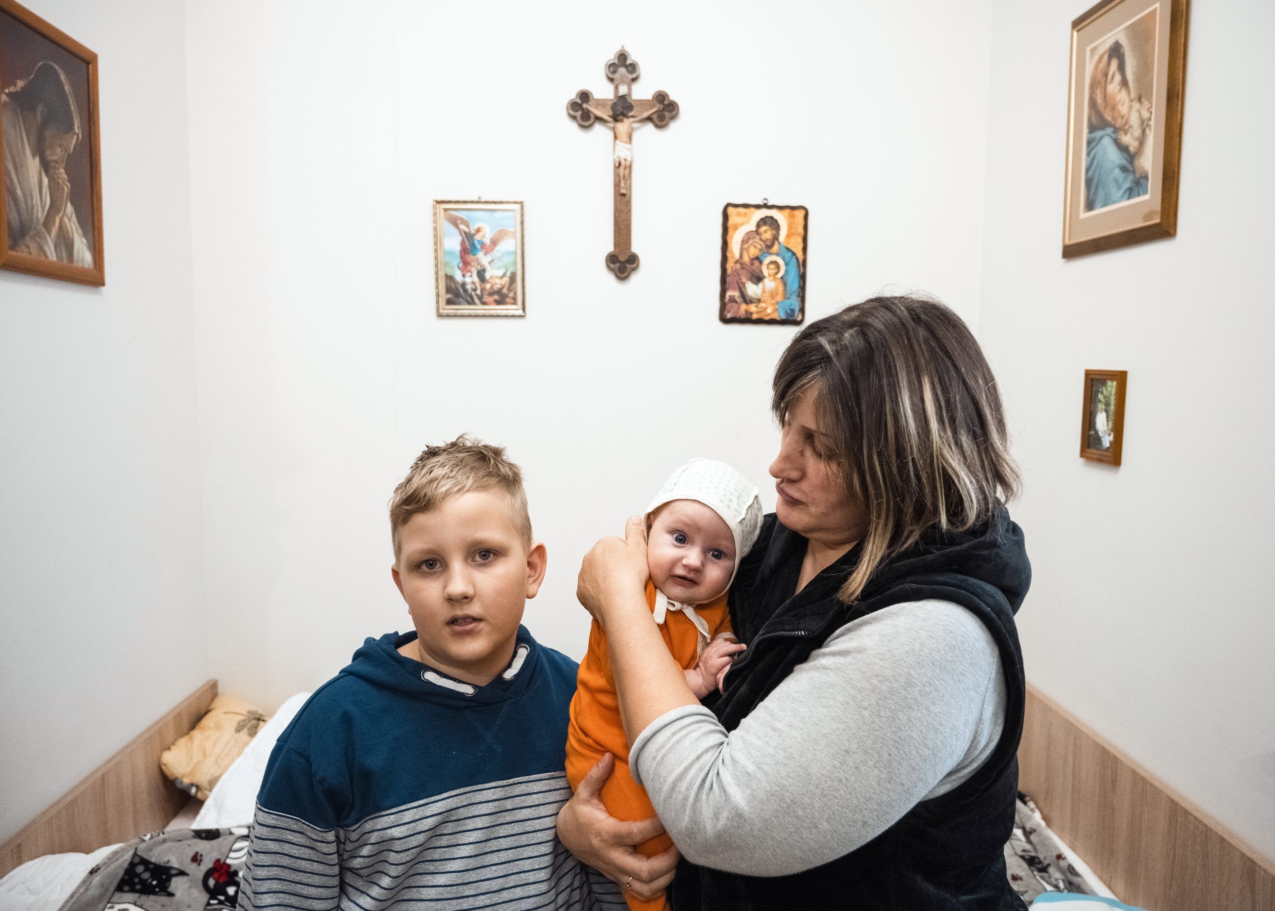 En mor holder sit lille barn i armene. Ved siden af står hendes søn.